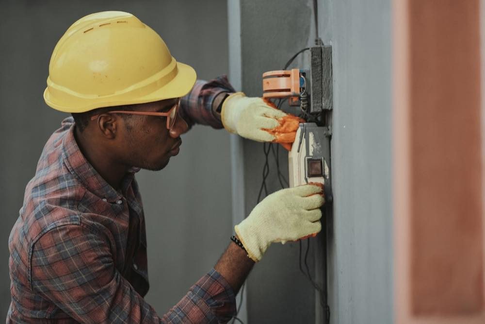 A tradesperson in a hard hat and gloves working on a wall-mounted electrical box.